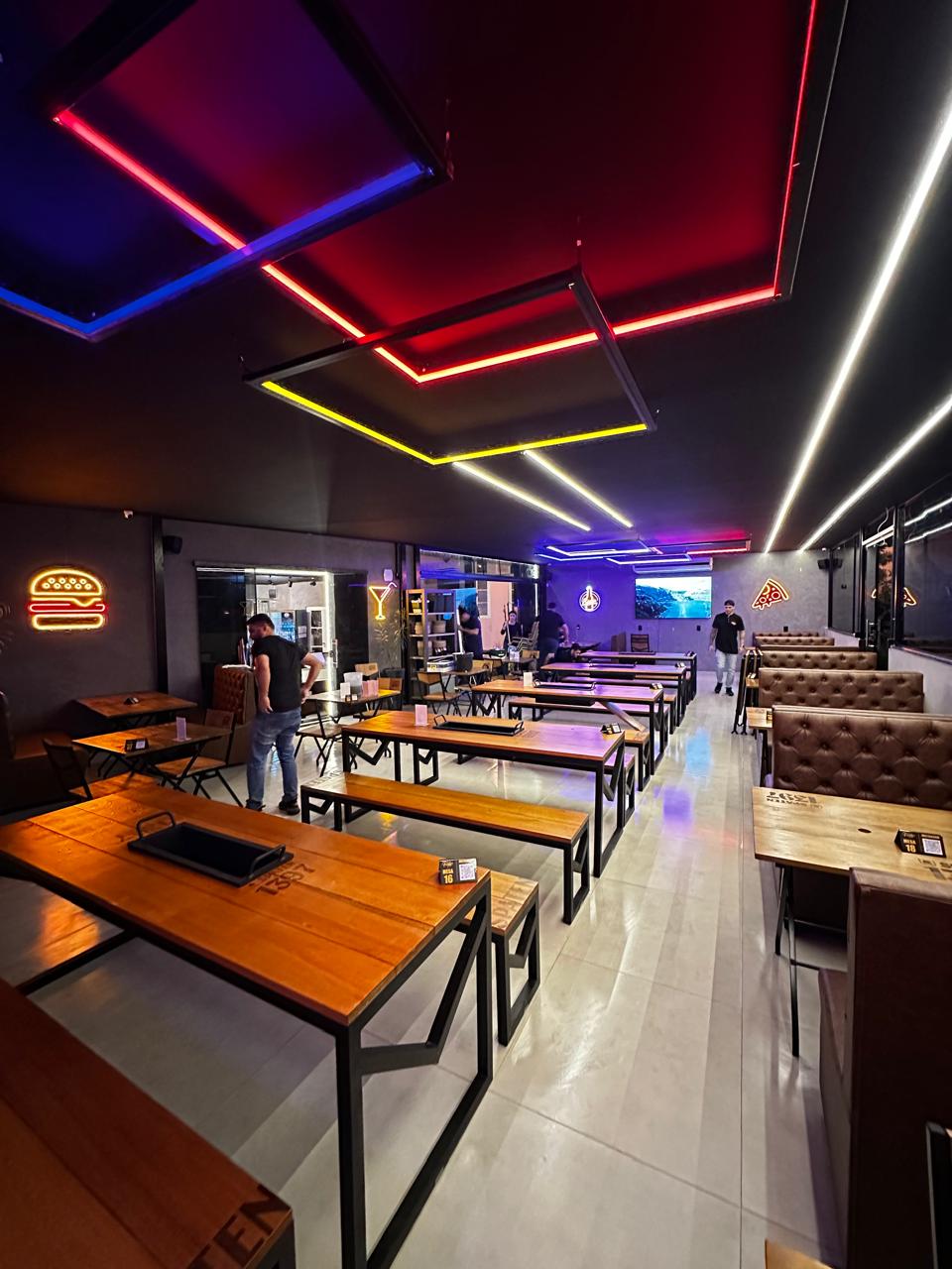 Interior do The Family's Sorocaba com iluminacao neon colorida, mesas de madeira e sofas estofados